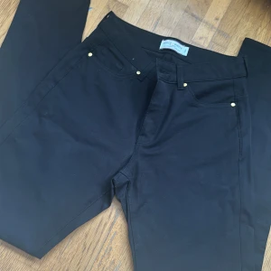 Svarta byxor från Holly & Whyte - Snygga svarta byxor från Holly & Whyte med dragkedja och knappar i guld. Perfekta för en stilren look. Byxorna har en klassisk design med fem fickor och är tillverkade i ett bekvämt material.