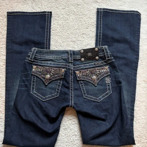 Miss Me jeans - Säljer ett par mörkblå, lågmidjade jeans från Miss Me med snygga broderade och dekorerade bakfickor. Jeansen har vita kontrastsömmar, bootcut och detaljerade nitar. Innerbenslängd: 86 cm. Kontakta mig för fler bilder eller vid intresse!💕