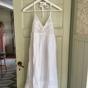 Vit klänning med virkad överdel - Superfin vit klänning med virkad överdel och knytband i nacken. Kjolen är luftig och faller lätt, perfekt för varma dagar. Klänningen har en bohemisk känsla och är ärmlös med en lätt rynkad nederdel.