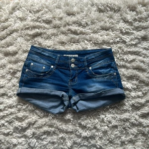 Lågmidjade shorts - Snygga blå jeansshorts med dubbla knappar och fickor fram och bak. Shortsen har uppvikta ben och kontrastsömmar som ger en cool look. Perfekta för varma dagar och enkel att matcha med olika toppar.