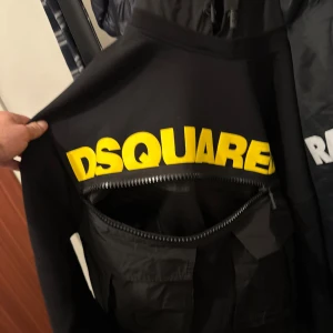 Svart hoodie från Dsquared2 med gul logga - Säljer en svart hoodie från Dsquared2 i storlek M. Tröjan har en stor gul DSQUARED2-logga på ryggen och en unik dragkedjedetalj. Materialet är mjukt och bekvämt, perfekt för dig som gillar streetwear och vill sticka ut.
