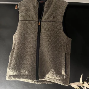Grå fleeceväst med dragkedja. Med märket Morris  - Morris grön fleeceväst med hel dragkedja framtill och broderad detalj på bröstet. Västen har en normal passform och är perfekt för lager-på-lager under kyliga dagar. Enkel och stilren design som passar till många outfits.