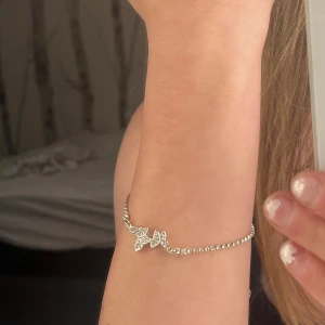 Silverfärgat glittrit armband med fjärilar  - Säljer ett elegant armband i silverfärg med gnistrande strass och två dekorativa fjärilar framtill. Armbandet har justerbar kedja för perfekt passform och ger en lyxig känsla till din stil. 