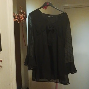 Svart blusklänning med rosett från Boohoo - Säljer en svart blusklänning från Boohoo med transparenta, prickiga ärmar och volangdetaljer vid ärmslut. Klänningen har en söt rosett framtill och är tillverkad i ett lätt, syntetiskt material. Perfekt för dig som gillar en feminin och trendig stil.