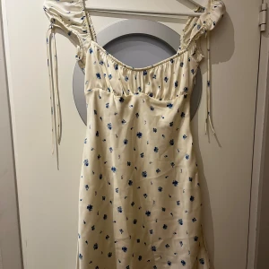 Blommig ljus klänning från Zara - Supersöt ljus klänning från Zara med små blå blommor. Klänningen har puffiga korta ärmar med knytband och en smickrande rynkad byst. Perfekt för sommarens alla tillfällen! Endast använd 1 gång och säljer då jag upplever den lite för kort på mig som är 175,5 cm lång 