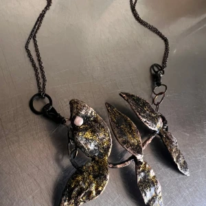 Unikt handgjort halsband med bladformade hängen - Säljer ett handgjort halsband med stora, bladformade hängen i metall. Hängena har en rå, konstnärlig finish med inslag av svart och guld, och sitter på en mörk kedja. Perfekt för dig som gillar statement-smycken och unika accessoarer.