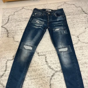 DNM Oscar jeans  - Säljer ett par mörkblå jeans med slitningar och patch-detaljer på framsidan samt små färgstänk i olika färger. Klassisk femficksmodell med knapp- och dragkedjegylf. Perfekta för en trendig och avslappnad look.
