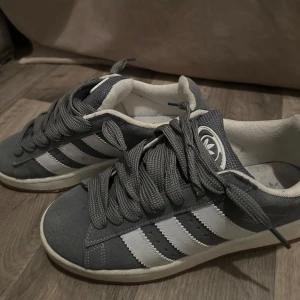 Grå Adidas Campus sneakers - Säljer ett par grå Adidas Campus sneakers med vita ränder och vit sula. Skorna har breda gråa skosnören och klassisk Adidas-logga på plösen. Ovandelen är i mocka och insidan är vit. Perfekta för en stilren vardagslook.