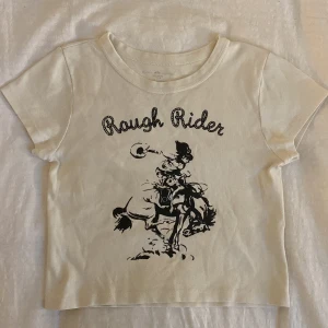 Vit t-shirt med cowboy-tryck från Brandy Melville - Vit kort t-shirt från Brandy Melville med svart tryck och texten 'Rough Rider' på bröstet. Klassisk rund hals och korta ärmar. Är mindre i storlek och mer som en baby-tee än vad som syns på bilderna, har några knappast synbara fläckar som man inte märker i verkligheten 🫶 passar S och vitare i verkligheten.
