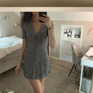 Blommig kortklänning från BikBok - Supersöt kortklänning från BikBok med blommigt mönster i vitt och blått på mörk botten. Klänningen har v-ringning, korta ärmar och knappar framtill. Perfekt för sommarens alla tillfällen!