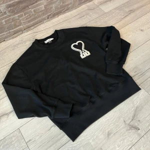 Svart ami paris hoodie - Säljer en svart sweatshirt från Ami Paris med deras ikoniska hjärtformade A-logga i vitt på bröstet. Tröjan har rund halsringning och en klassisk, avslappnad passform. Perfekt för en stilren och trendig look.
