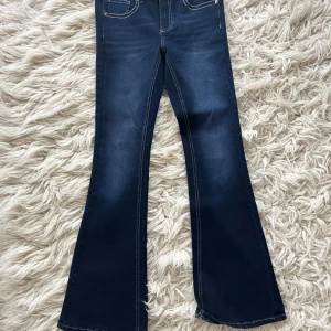 Mörkblå low waist bootcut jeans med dekorficka - Helt nya mörkblå bootcut jeans från RYVLS  med kontrastsömmar och dekorativ ficka med knapp och nitar bak. Klassisk femficksmodell och low waist. Stl 164, innerbenslängd 80 cm. 
