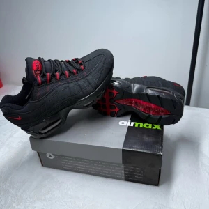 Nike Air Max 95 Svarta Sneakers med Röda Detaljer - Säljer ett par svarta Nike Air Max 95 sneakers med röda detaljer och snörning. Skorna har en sportig design med synlig Air Max-dämpning i sulan och mesh- och syntetmaterial på ovandelen. Perfekta för dig som gillar streetwear och vill sticka ut.