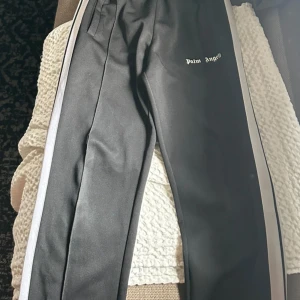 Svarta byxor med vit sidoremsa från Palm Angels - Säljer ett par svarta byxor från Palm Angels med vit sidoremsa längs benen och logga tryckt på ena låret. Byxorna har resår i midjan och raka ben, perfekt för en sportig och trendig look.