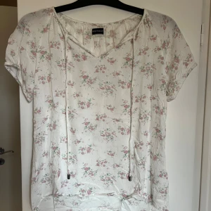 Blommig kortärmad blus från Miss Milla - Söt blommig blus från Miss Milla i vitt med små rosa och gröna blommor. Blusen har korta ärmar, v-ringning och knytband vid halsen. Perfekt för vår och sommar!