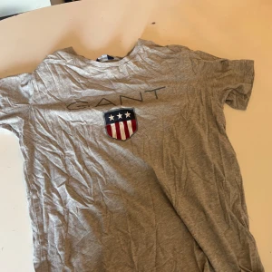 Grå t-shirt från GANT - Säljer en grå t-shirt från GANT med ett tryck av en sköld med stjärnor och ränder på framsidan. T-shirten har korta ärmar och rund halsringning. Perfekt för en avslappnad stil.