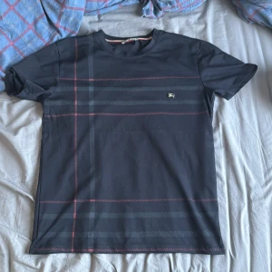 Burberry T-shirt - Snygg svart t-shirt med diskret rutigt mönster i rött och grått. Perfekt för en stilren look. Kortärmad och tillverkad i ett mjukt material för extra komfort.
