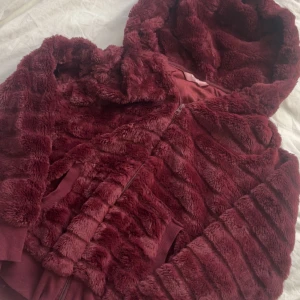 pälsjacka  - Mysig vinröd pälsjacka från hunkmöller med huva och dragkedja. Jackan har en fluffig textur och ribbade muddar vid ärmsluten. Perfekt för kyliga dagar när du vill hålla dig varm och stilren. 