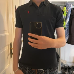 Mörkblå pikétröja från Polo Ralph Lauren - Säljer en klassisk mörkblå pikétröja från Polo Ralph Lauren med röd broderad logga på bröstet. Tröjan har korta ärmar, krage och knappar framtill. Perfekt för en stilren och avslappnad look. Passar både S och Xs! Nypris ca 1000kr
