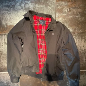 Svart bomberjacka från Fred Perry - Klassisk svart bomberjacka från Fred Perry med röd- och vitrutigt foder. Jackan har dragkedja framtill, ribbade muddar och krage. Perfekt för dig som vill ha en stilren och tidlös look.
