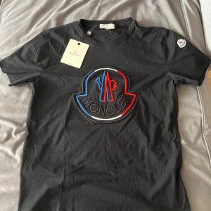 Svart Moncler t-shirt med stor logga - Säljer en svart Moncler t-shirt med en stor logga i blått, rött och vitt på bröstet. T-shirten är oanvänd och i mycket bra skick. Storlek S men även stor i storleken så passar även M.