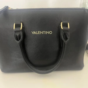 Svart handväska från Valentino - Snygg svart handväska från Valentino med guldfärgade detaljer och dubbla handtag. Väskan har dragkedja upptill och ett rymligt huvudfack med innerficka. Perfekt accessoar för att lyfta din outfit.