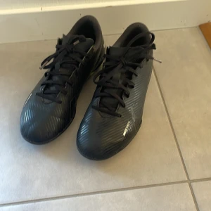 Svarta fotbollsskor från Nike - Säljer ett par svarta Nike Mercurial fotbollsskor med snörning. Skorna har en stilren design med diskreta vita detaljer och är perfekta för fotbollsplanen.