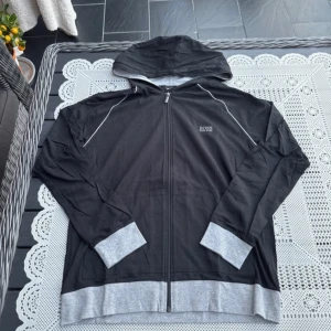 Svart och grå hoodie från Boss - Säljer en stilren svart hoodie från Boss med gråa detaljer. Den har en dragkedja framtill och en praktisk huva. Perfekt för en avslappnad look.
