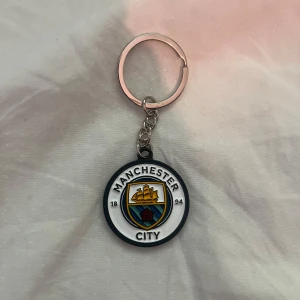 Manchester City nyckelring - Säljer en rund nyckelring med Manchester Citys klubbmärke. Nyckelringen har en metallkedja och själva märket är i plast med klubbens logga i blått, gult, svart och vitt. Perfekt för fotbollsfans!
