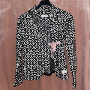 Svartvit omlottkofta med mönster och rosett - Säljer en svartvit omlottkofta från Odd Molly med grafiskt mönster och en ljusrosa rosett i midjan. Koftan har långa ärmar och är perfekt för dig som vill ha något både snyggt och bekvämt. Är i storlek 1 så motsvarar en S