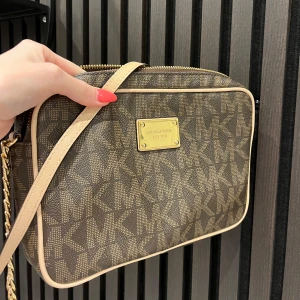 Brun axelväska från Michael Kors - Snygg brun axelväska från Michael Kors med guldiga detaljer och kedja på axelremmen. Väskan har MK-logga över hela ytan, dragkedja upptill och en platta med logotyp framtill. Perfekt storlek för det viktigaste och med ljus insida.