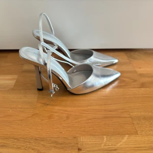 Silvriga pumps med klack och spetsig tå - Säljer ett par silvriga pumps från Bershka med hög klack och spetsig tå. Skorna har en smal rem runt hälen och öppen baksida. Perfekta för dig som vill sticka ut med metallic-look.