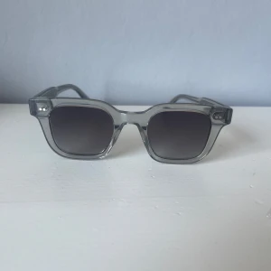 Grå transparenta solglasögon från Chimi - Stilrena solglasögon från Chimi i transparent grå plast med fyrkantig form och mörka glas. Snygg detalj med glittrig insida på skalmarna. Perfekta för att lyfta din look med en modern touch.