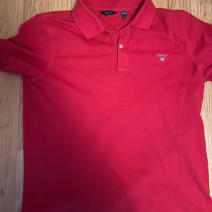 Röd kortärmad pikétröja från Gant - Säljer en snygg röd pikétröja från Gant med klassisk krage och knappdetalj framtill. Tröjan har korta ärmar och en liten broderad logga på bröstet. Perfekt för en stilren look!