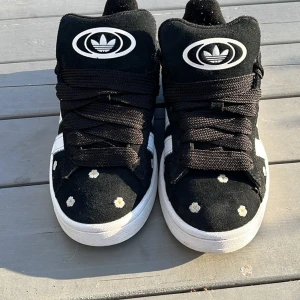 Svarta Adidas Campus med blommönster - Snygga svarta Adidas Campus sneakers med vita detaljer och ett charmigt blommönster. Skorna har klassiska tre ränder på sidan och en rund tå. Perfekta för en stilren look med en twist.