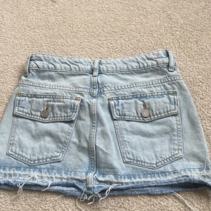 Ljusblå jeanskjol med rå kant - Säljer en ljusblå jeanskjol med klassiska fickor både fram och bak samt rå, fransig nederkant. Kjolen har knapp och dragkedja framtill och är i en kort modell. Perfekt för dig som gillar en avslappnad och trendig stil. Köpt i Spanien på pull and bear