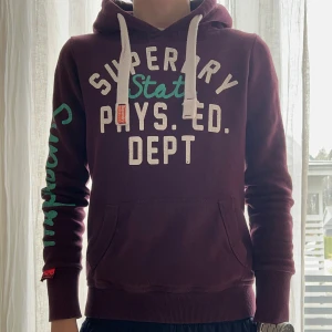 Vinröd hoodie från Superdry - Säljer en snygg vinröd hoodie från Superdry med texttryck i vitt och turkost. Tröjan har en klassisk känguruficka och justerbar huva med vita snören. Perfekt för en avslappnad stil. Väldigt stilren! Skönt snygg och skön, har krympt i tvätten. Och sitter liten på mig
