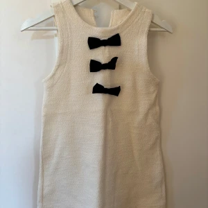 Vit ärmlös klänning med svarta rosetter från ZARA i storlek 10 /140 - Säljer en stilren vit ärmlös klänning med tre svarta rosetter framtill. Perfekt för en elegant look. Klänningen har en enkel design med en knapp i nacken för enkel på- och avtagning.