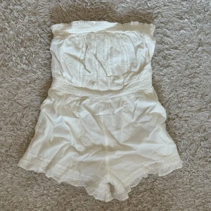 Vit axelbandslös jumpsuit - Säljer en söt vit axelbandslös jumpsuit med elastisk midja och spetsdetaljer. Perfekt för varma sommardagar. Från Bershka, ny med prislapp kvar. 
