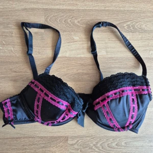 Svart och rosa bh från La Senza 80A - Säljer en snygg svart bh från La Senza med rosa spetsdetaljer. Bh:n har justerbara axelband och en dekorativ rosett framtill. Den har uttagbara inlägg. Finns några tecken på användning, se bilder.