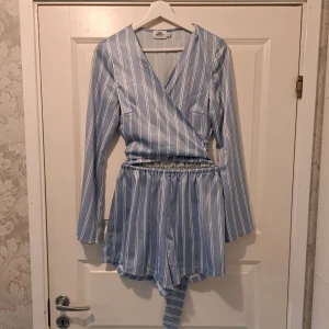 Blå randig omlottklänning från NA-KD - Snygg blå omlottjumpsuit från NA-KD designad av Andrea Hedenstedt. Dressen har ett randigt mönster och långa ärmar. Den har en knytning i midjan för en smickrande passform. Framsida visar lite mage. Perfekt för en stilren look!