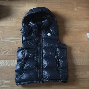 Blå dunväst från Moncler - Snygg svart dunväst från Moncler med dragkedja och huva. Perfekt för kyliga dagar. Västen har en quiltad design och en ikonisk Moncler-logga på bröstet. Insidan är fodrad i en mörkare nyans med en rolig serietidningsdetalj.