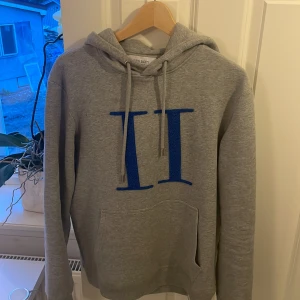 Grå hoodie från Les Deux - Säljer en stilren grå hoodie från Les Deux med en stor blå romersk siffra på framsidan. Tröjan har en klassisk känguruficka och justerbar huva med snören. Perfekt för en avslappnad stil.