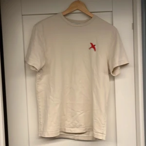 Beige t-shirt från Axel Arigato - Säljer en stilren beige t-shirt från Axel Arigato med en liten röd broderad detalj på bröstet. T-shirten är kortärmad och gjord i mjukt material, perfekt för en avslappnad look.