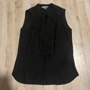 Svart ärmlös blus från H&M - Snygg svart ärmlös blus från H&M med en elegant volangdetalj framtill. Perfekt för en stilren look, oavsett tillfälle. Blusen har en klassisk passform och är tillverkad i ett lätt och bekvämt material.