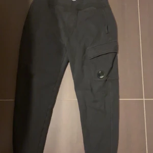 Svarta cargopants från C.P. Company - Snygga svarta cargopants från C.P. Company med en praktisk ficka på sidan och knappdetalj. Perfekta för en avslappnad stil med en modern touch. Byxorna har en bekväm passform och är tillverkade i ett slitstarkt material. Redo för prisändring vid snabb affär?