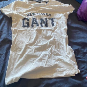 Vit t-shirt från GANT - Säljer en vit t-shirt från GANT med texten 'NEW HAVEN GANT' tryckt på framsidan. T-shirten har en rund halsringning och korta ärmar. Perfekt för en avslappnad stil. 🕶️storlek xs passar bra till barn runt 10-13 år. Nästan helt oanvänd då jag fick den i födelsedagspresent för flera år sen och den var för liten.