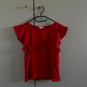 Röd topp med spetsdetaljer - Säljer en röd topp med spetsdetaljer på ärmarna. Toppen har en lös passform och korta ärmar med volang. Perfekt för en somrig look!