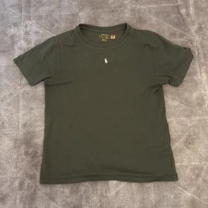 Grön t-shirt från Ralph Lauren - Säljer en stilren grön t-shirt från Ralph Lauren med en liten broderad logga på bröstet. T-shirten är kortärmad och gjord i mjuk bomull. Den har storlek 150 i barnstorlekar. Perfekt för en avslappnad look.