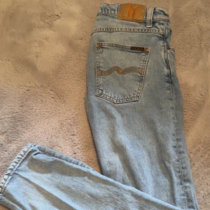 Ljusblå jeans från Nudie Jeans - Säljer ett par ljusblå jeans från Nudie Jeans med klassisk femficksdesign och dragkedja. Jeansen har en snygg broderad detalj på bakfickan och är i en straight passform. Perfekta för en avslappnad stil. Jeansen passar mig som oftast bär 29/30 -32